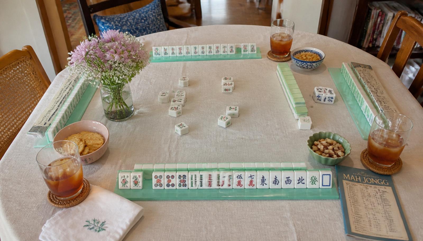 A casual, lived-in mahjong table set up at home with tiles mid-game, iced tea, and everyday snacks